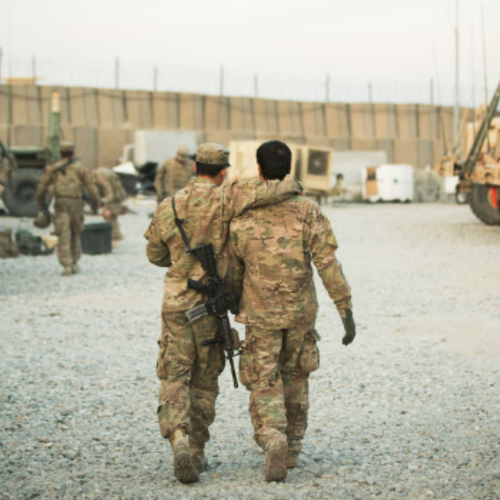 Two male soldiers in camouflage walk on a military base. The soldier on left has his a large gun slung around his back and his right arm wrapped around the other soldier's neck in a friendly embrace. Gravel is on the ground and a large wall is visible.